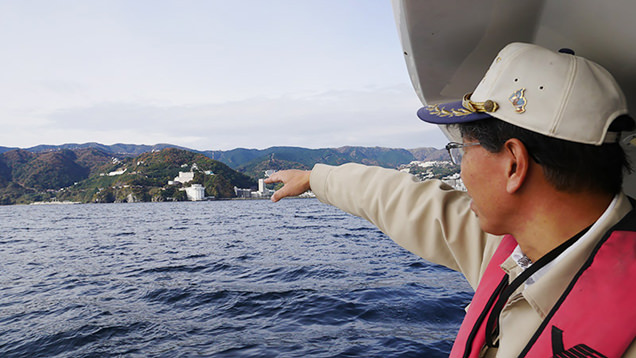 船に乗り向こうの島を指さす西海尊志さん